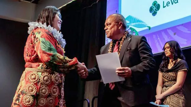 A woman dressed in a colourful traditional Pacific Island dress shakes hands with a man in formal attire while receiving a certificate on stage. Another woman stands nearby, also wearing traditional clothing, with a 'Le Va' logo displayed in the background.