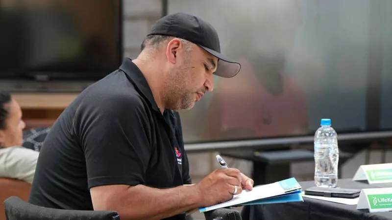 Man in black t-shirt and cap writing in workbook at a desk