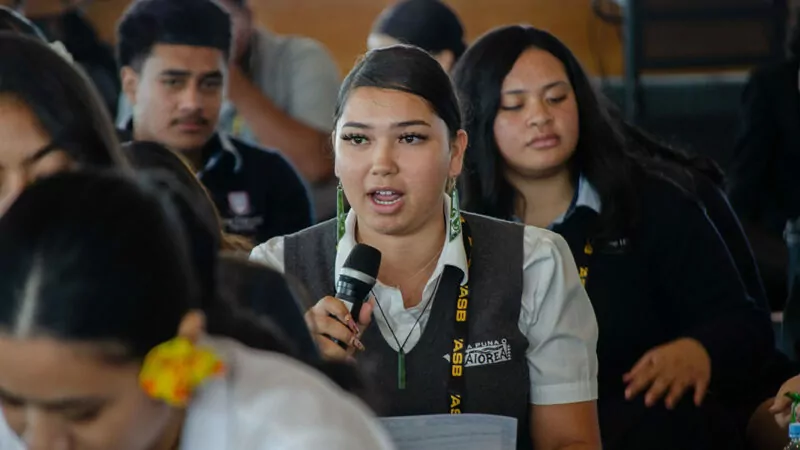 Young woman speaks into a microphone, seated among other students