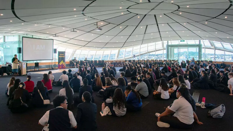 Large group of students sitting on the floor of a very big room, listening to a presenter