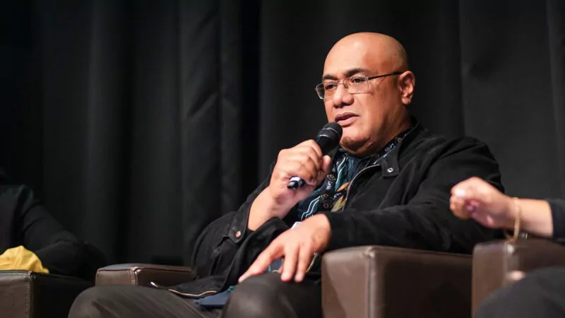 Manase Lua speaking into a microphone while seated in an armchair on stage