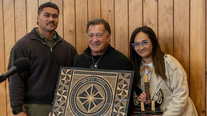 Older man holding a Pacific carving, with a young man on the left and young woman on the right.