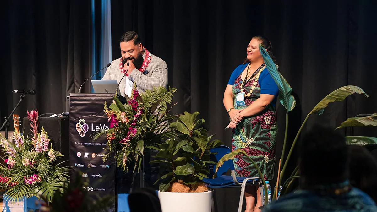 Ben Tameifuna and Sara-Jane Elika stand on a stage, smiling, with plants along the front of the stage